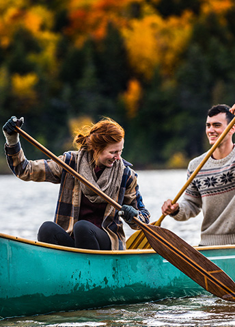 A smiling man and woman paddling a teal canoe on a lake surrounded by autumn trees.